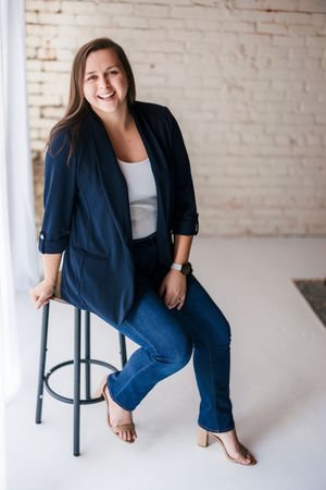 Professional wearing navy blazer and white top poses casually on bar stool against white brick wall in studio setting.