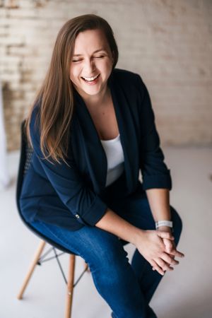 Casual portrait showing joyful laughter while seated in navy blazer and white top against neutral background.