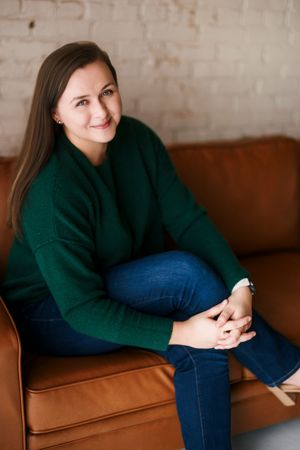 A person in a dark green sweater and blue jeans lounges comfortably on a brown leather couch.
