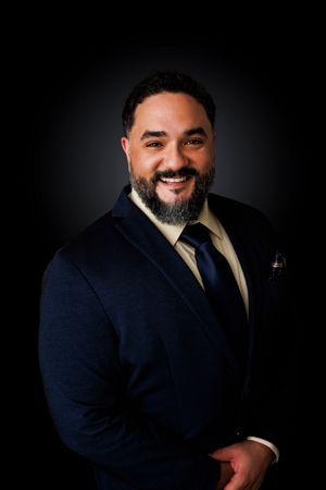 Professional headshot portrait of person in navy suit against dark background with dramatic lighting and warm smile.