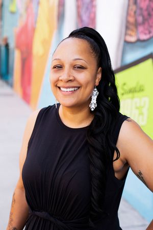 Smiling person wearing a black sleeveless dress and ornate silver earrings poses against colorful wall art background.