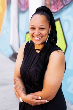 Woman in black sleeveless top poses confidently against colorful graffiti wall background.