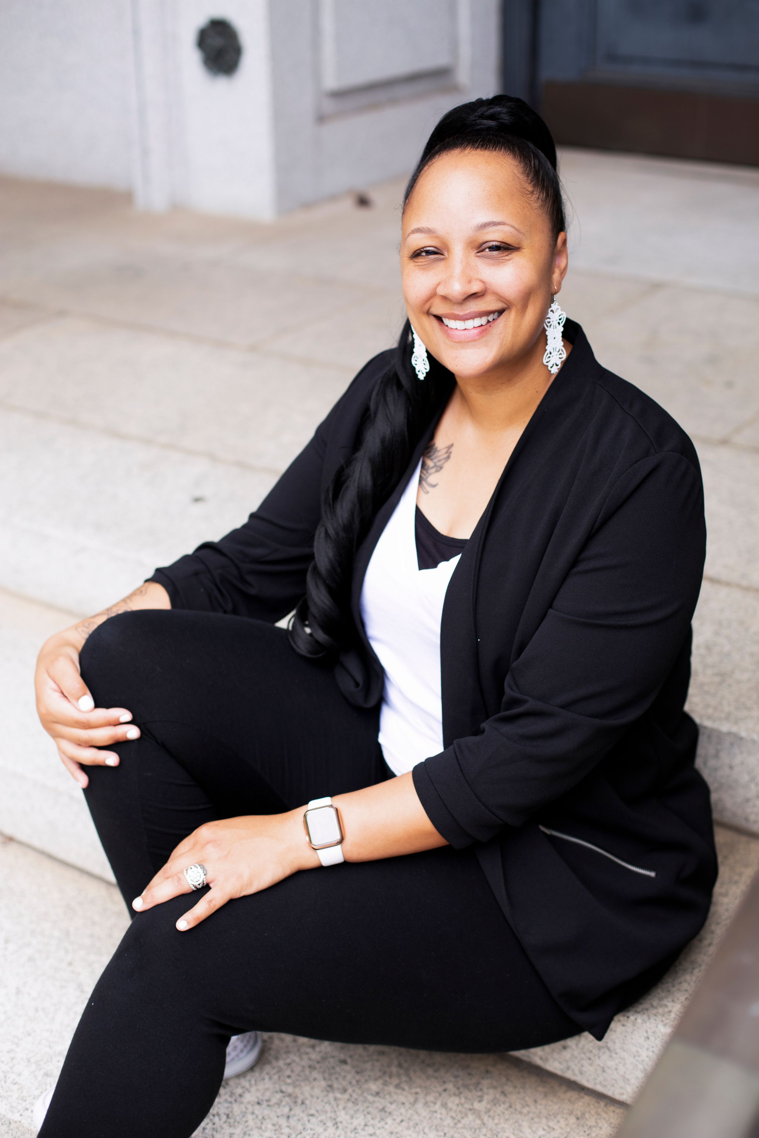 Professional portrait of someone in black and white business attire sitting on stone steps with a warm smile.