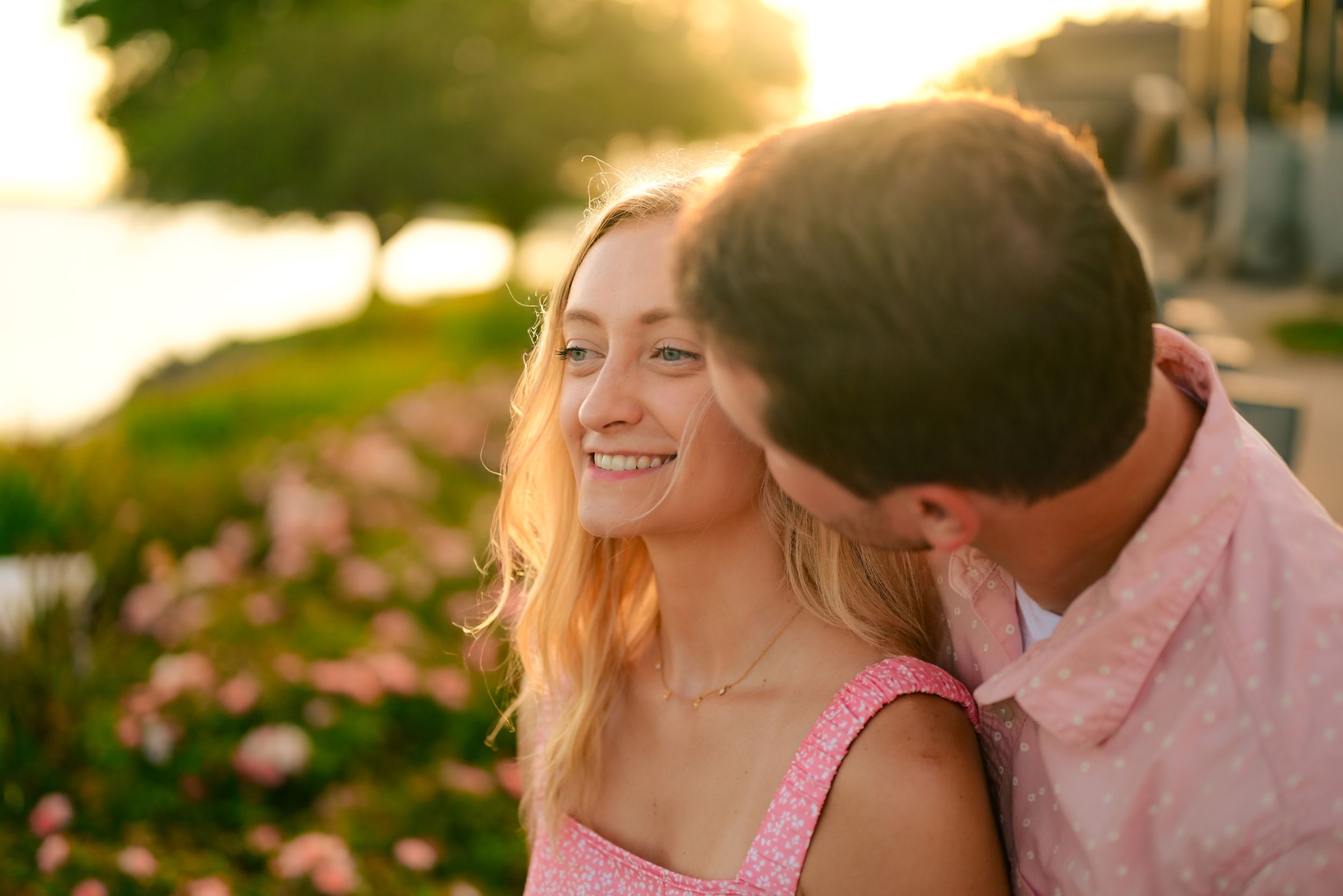 A couple in pink and white outfits share an intimate moment at sunset in a garden setting.