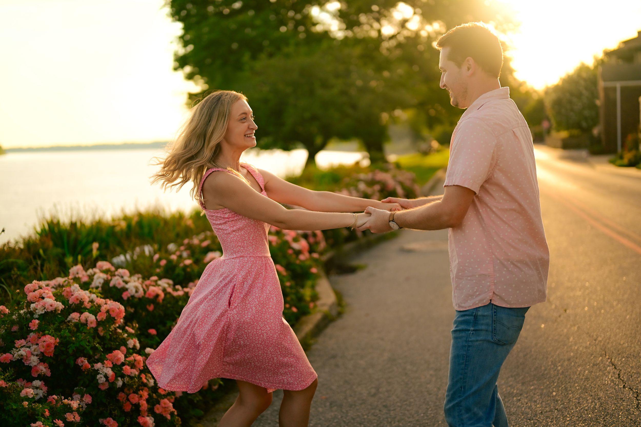 Couple dancing together on a path at sunset with golden light and pink flowers in the background.