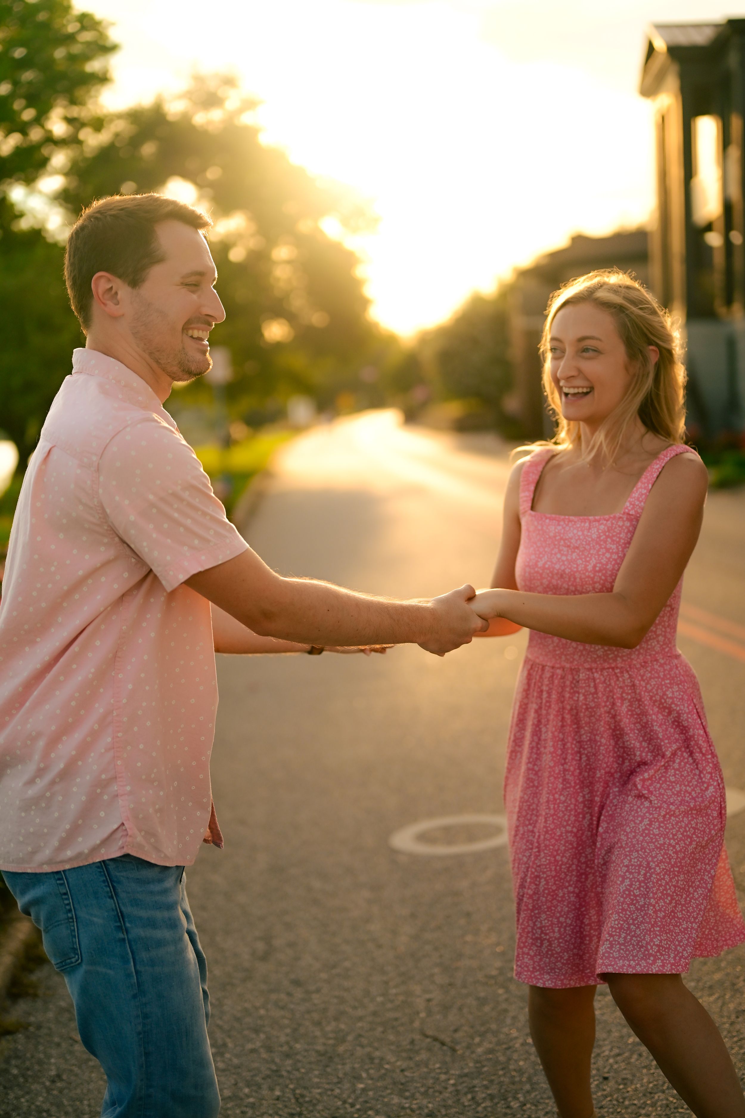 A couple dances playfully together at sunset on a path, with her wearing a pink dress and him in a light shirt.