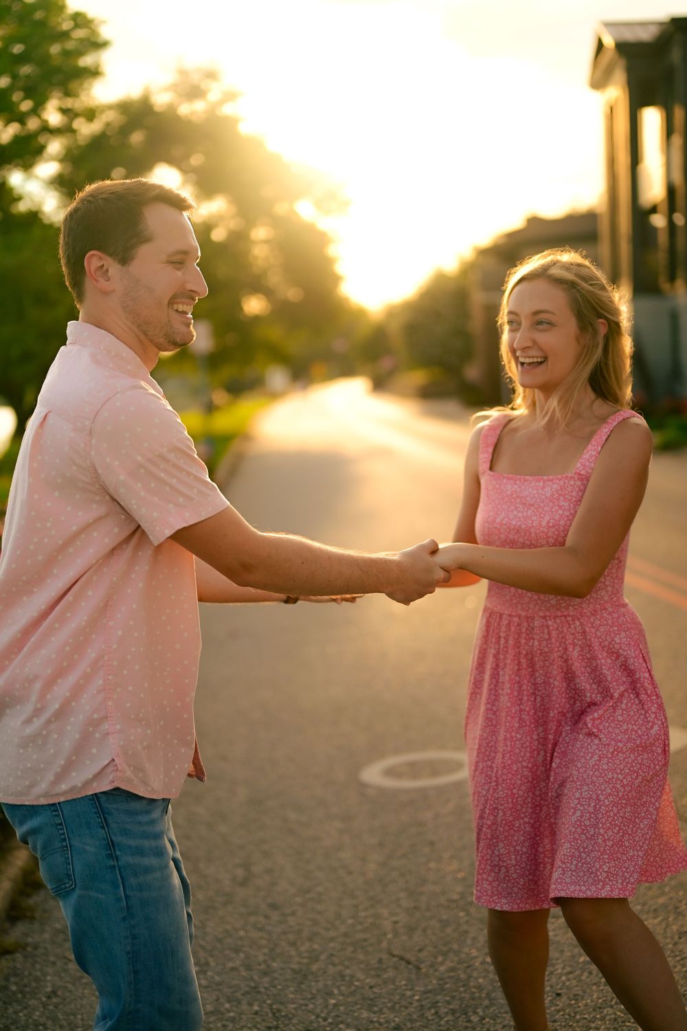 A couple dances playfully together at sunset on a path, with her wearing a pink dress and him in a light shirt.