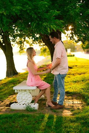 A romantic marriage proposal scene unfolds under green trees during golden hour sunset in a park setting.