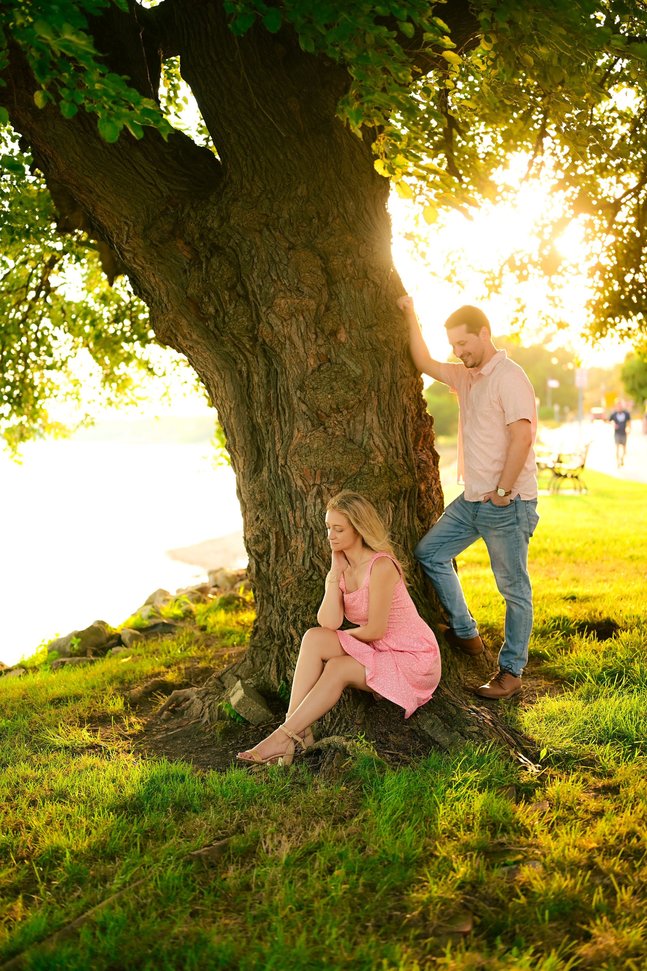 A couple sits together under a large tree at sunset in a grassy field during a romantic photoshoot.