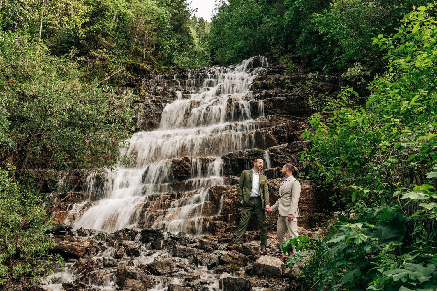 A cascading waterfall flows over rocky ledges surrounded by lush green forest in a scenic natural setting.