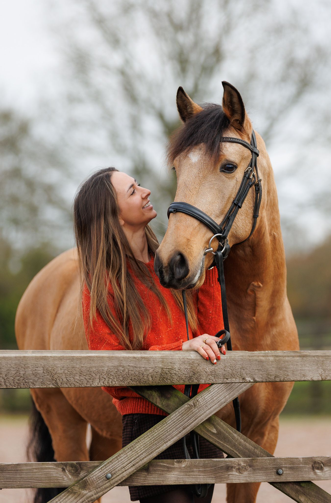 A person in a pink sweater and jeans stands with a black horse against a backdrop of golden autumn foliage.