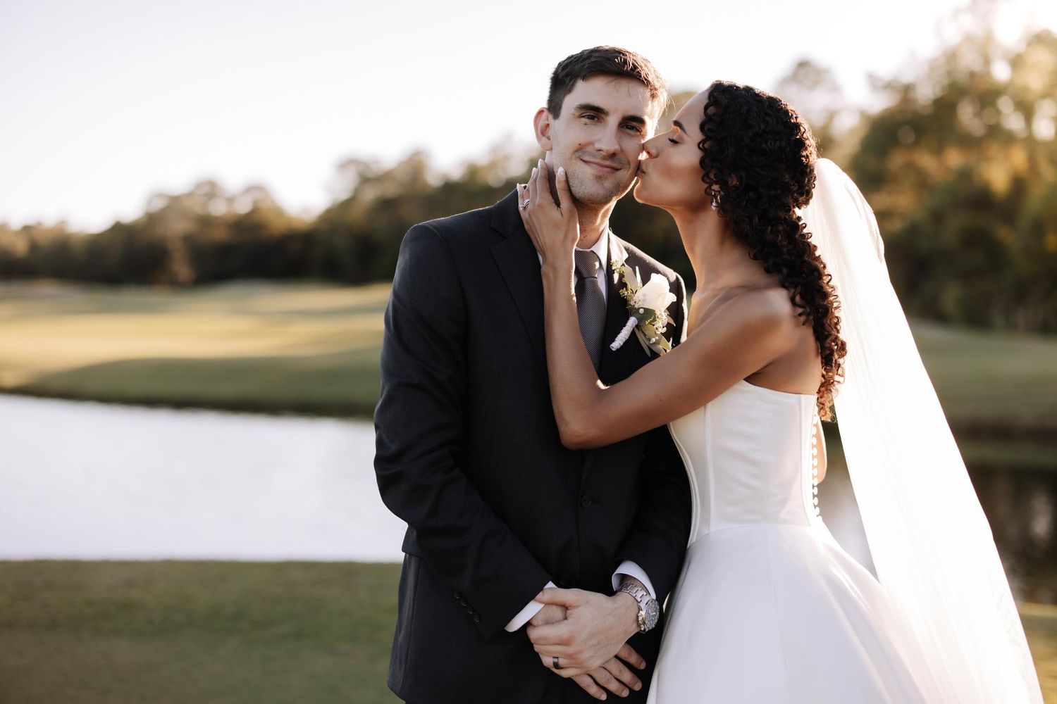 A romantic wedding couple embraces outdoors at sunset near a scenic lake and golf course.