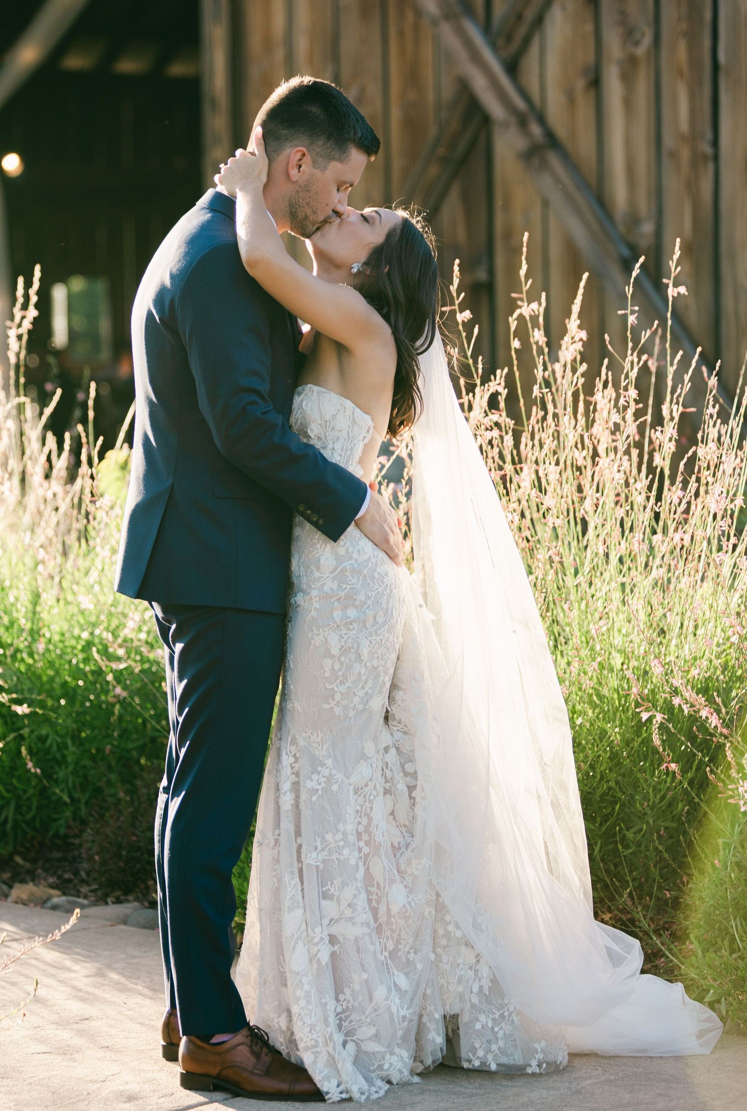 A romantic couple in wedding attire share intimate moments in a rustic outdoor setting with tall grass at sunset.