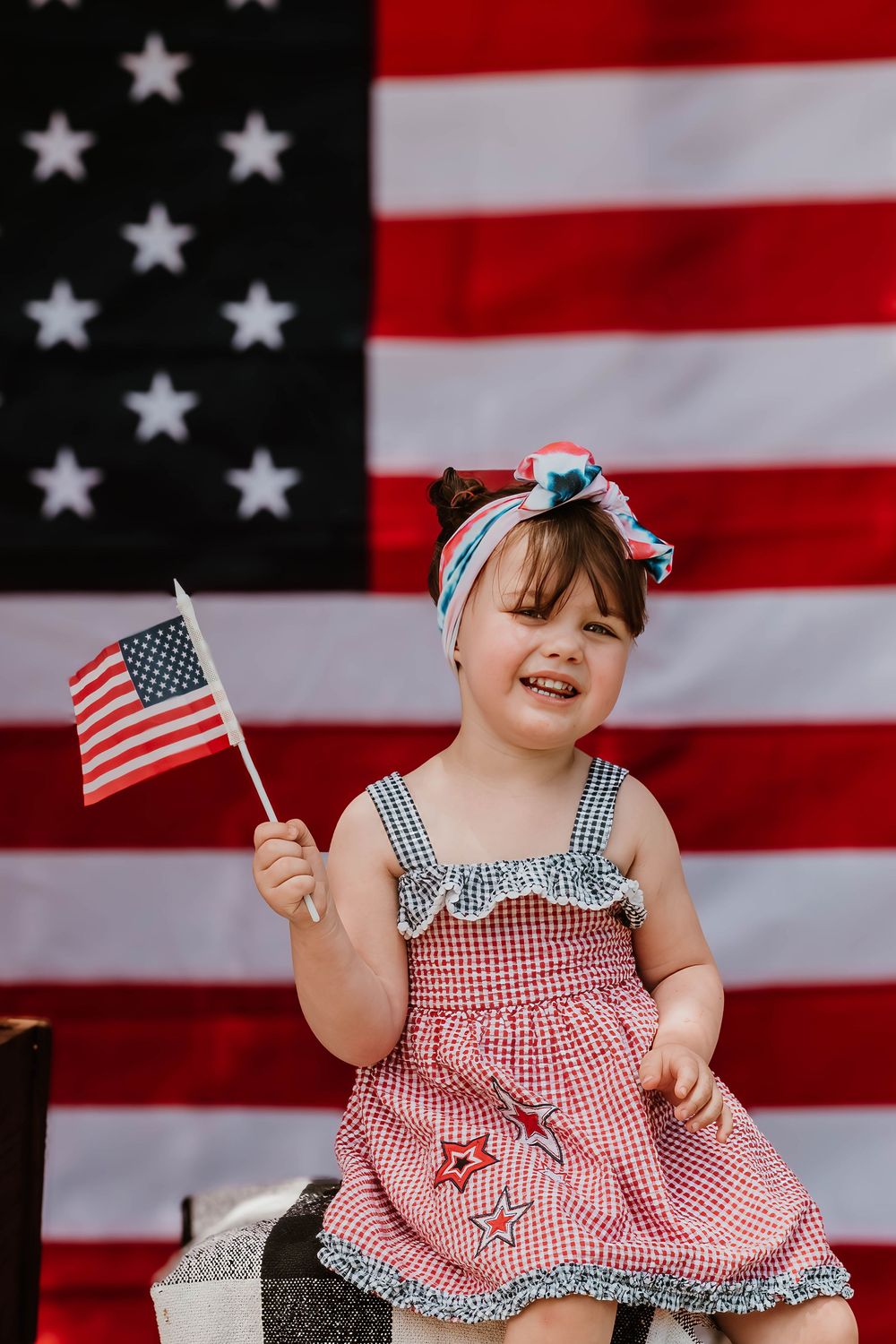 Young child in red gingham dress waves small American flag while sitting against large flag backdrop.