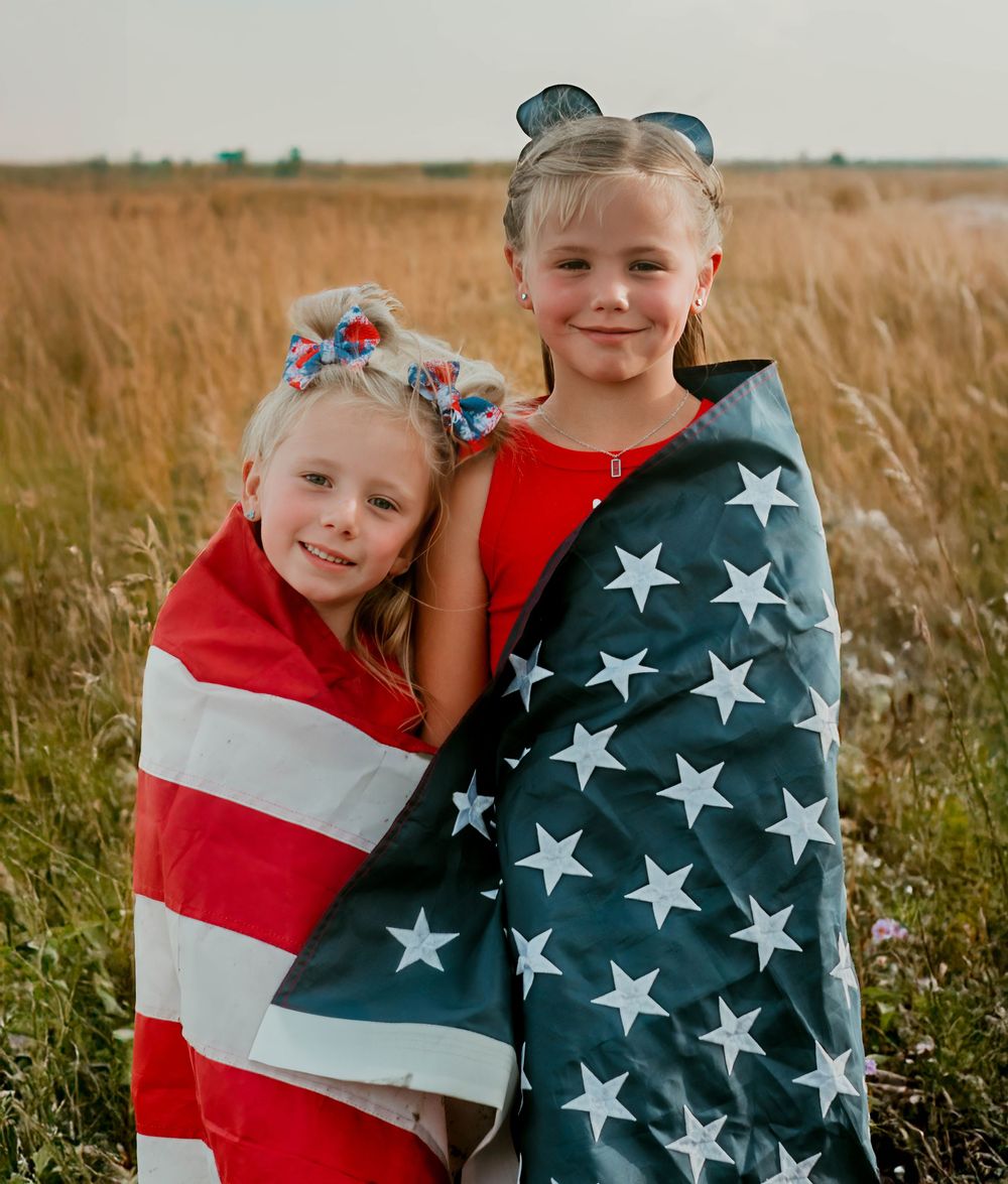 Children wrapped in American flag stand in golden wheat field at sunset.