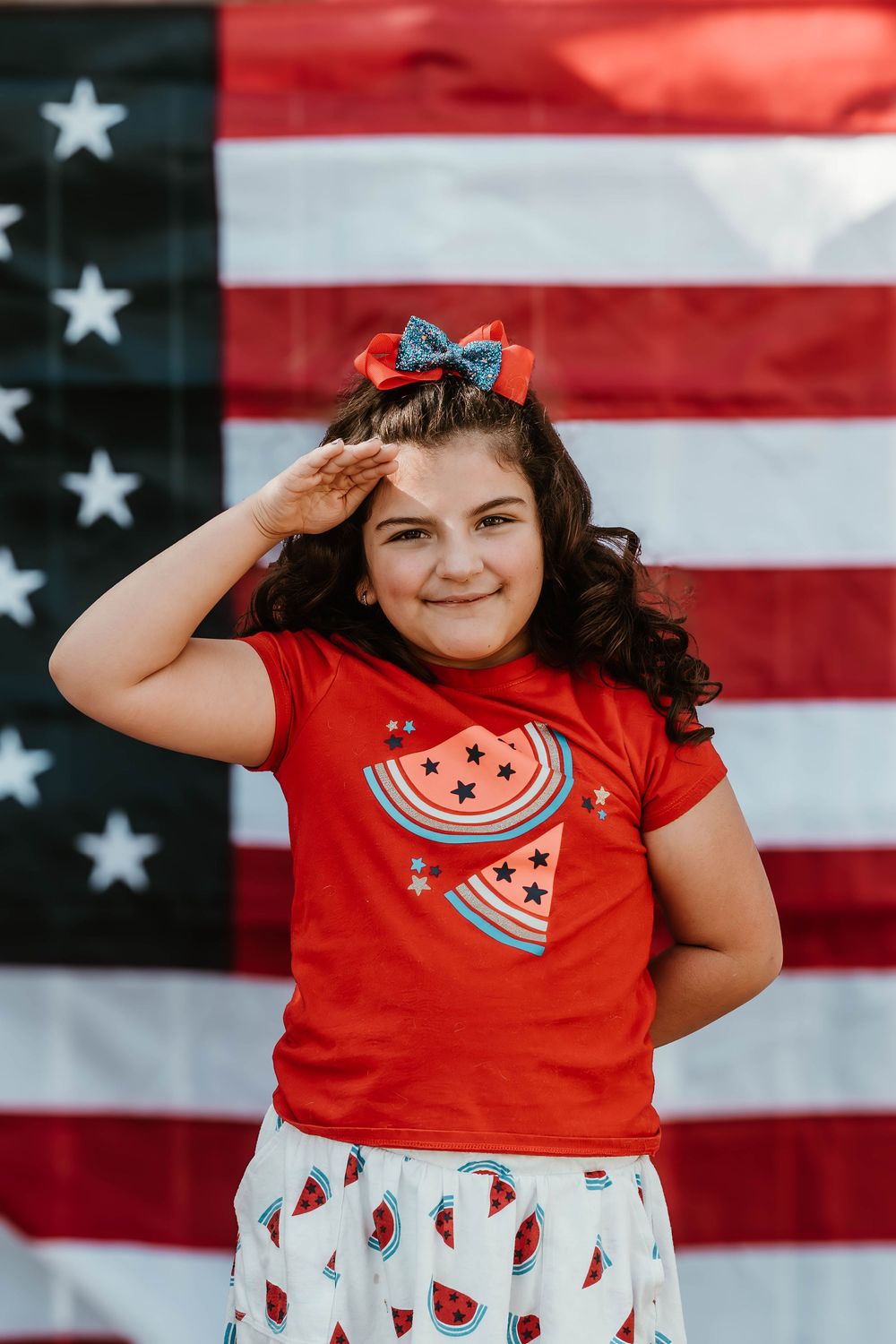 Child in red watermelon shirt salutes while posing against American flag backdrop.