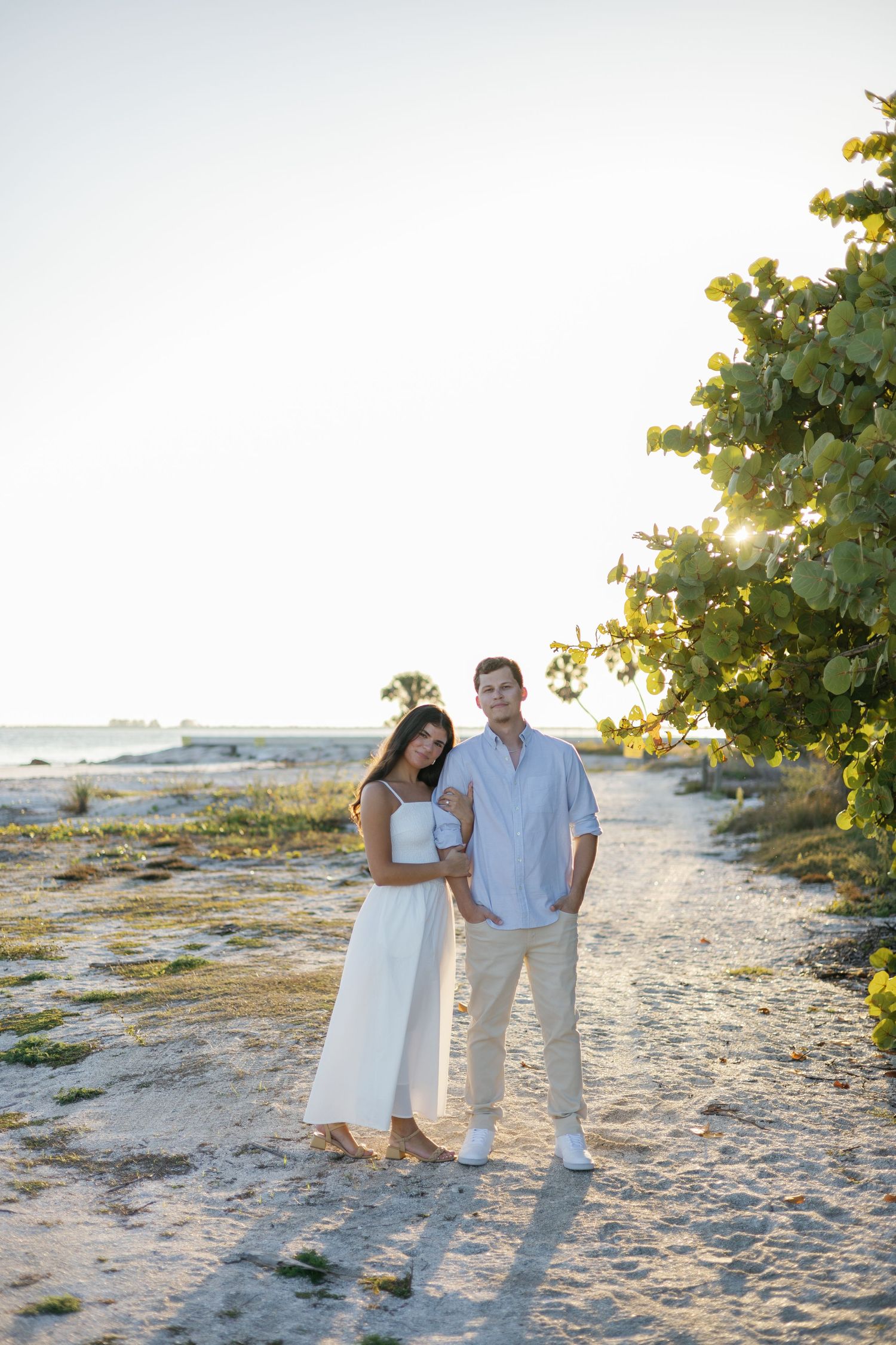 A romantic couple relaxes together on a white sandy beach during sunset with waves crashing in the background.