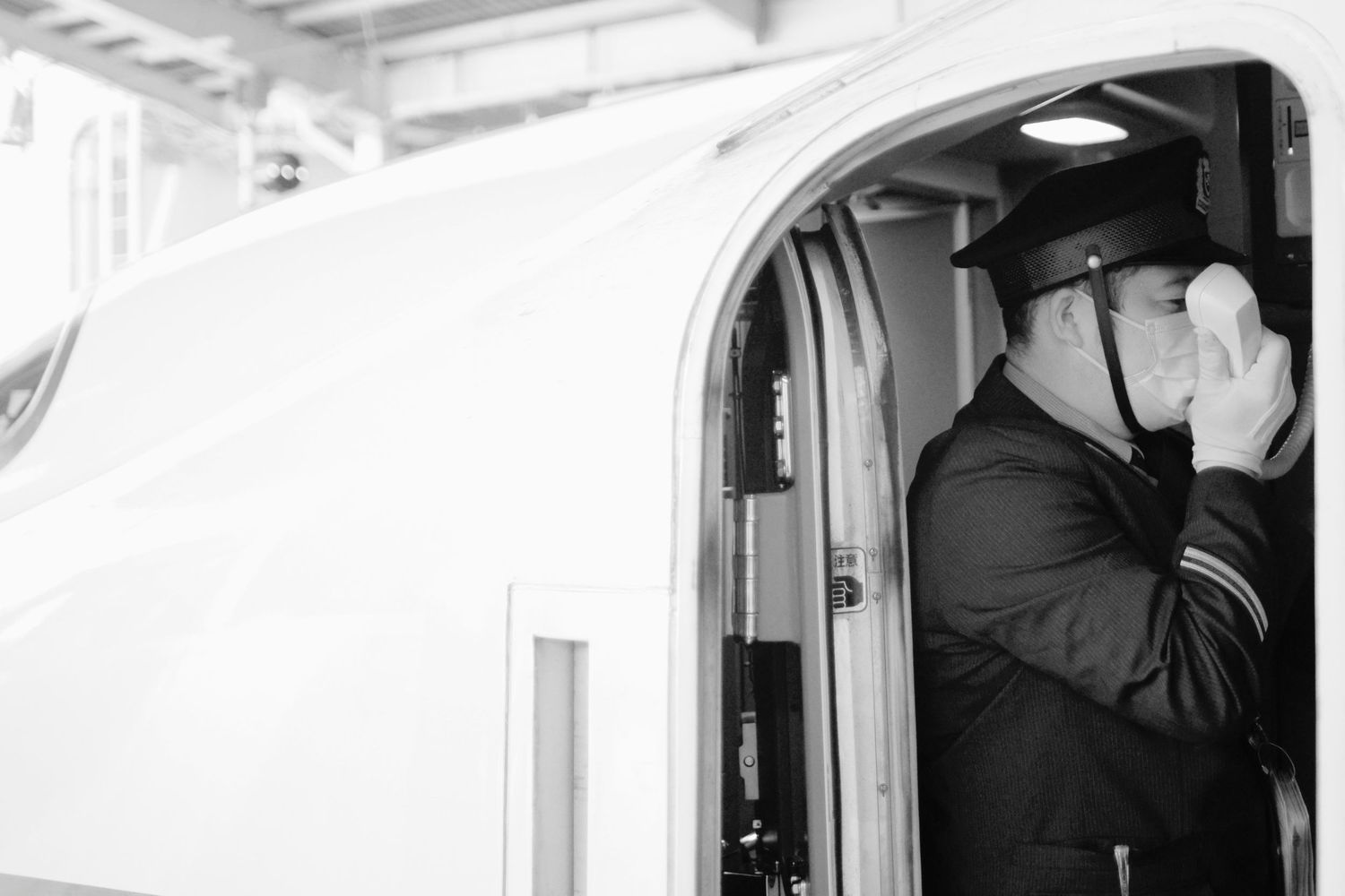 A train conductor in uniform peers through binoculars while standing in a doorway of a passenger car.
