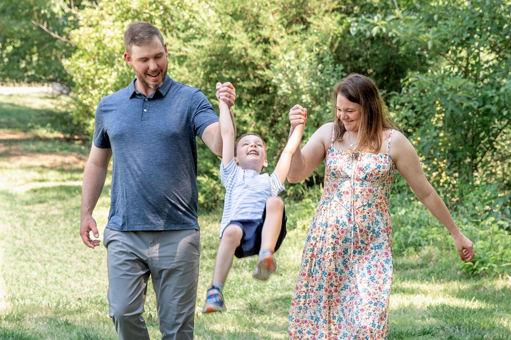 A happy family swings their young child between them outdoors in a lush green park on a sunny day.