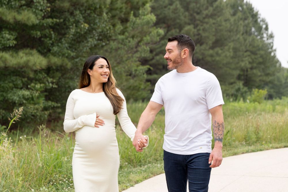 Pregnant woman in white dress holds hands with man in white tee, walking on a path surrounded by greenery.