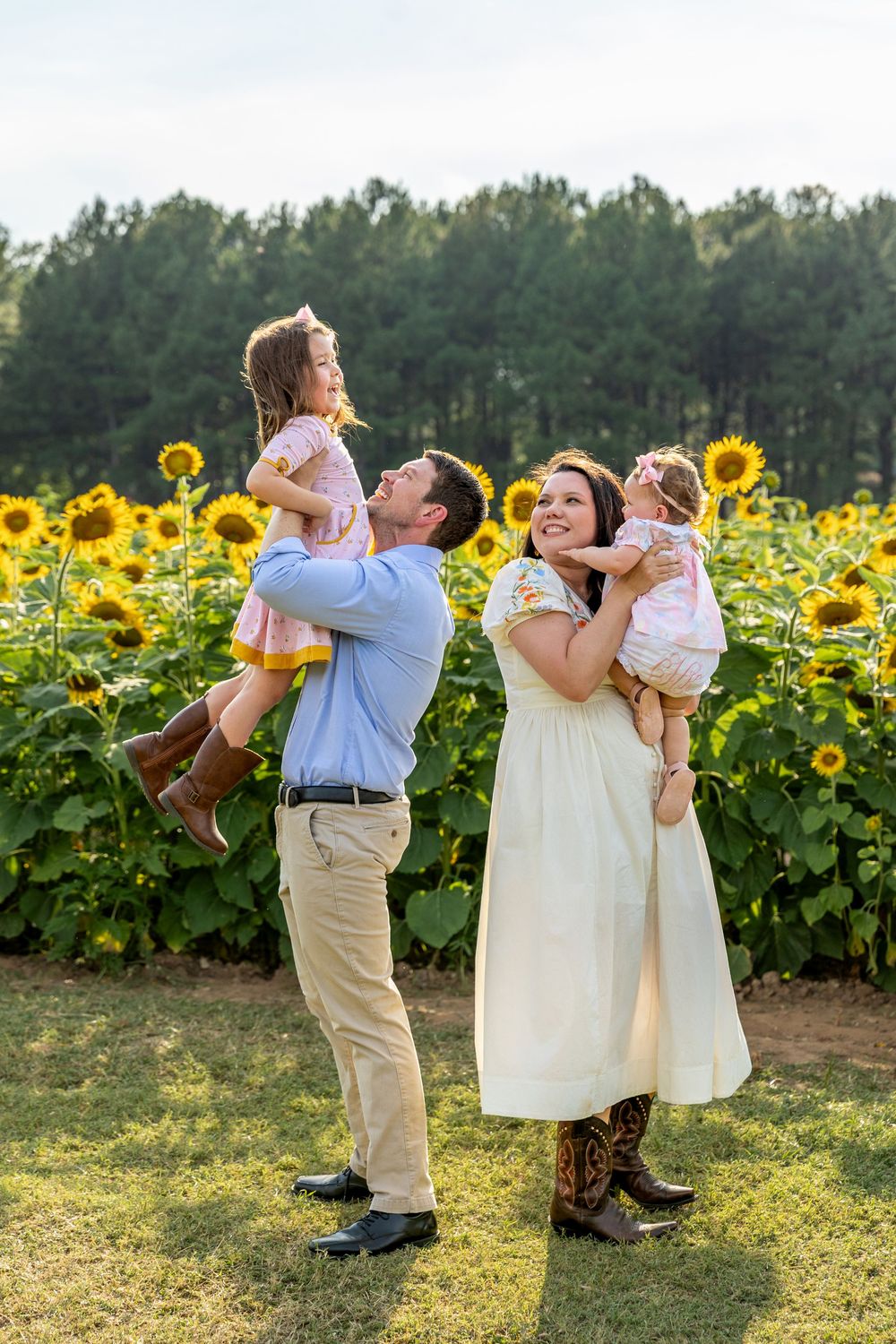 Family of four posing in a sunflower field, parents each holding a young child, smiling joyfully outdoors.