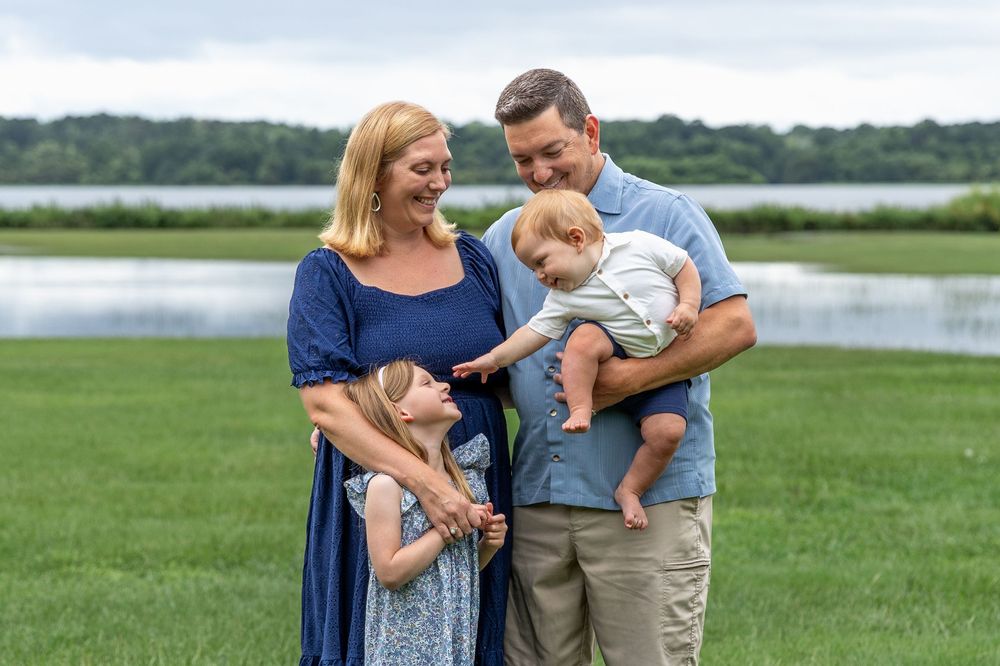 Happy family of four posing outdoors near a lake, parents smiling with young daughter and baby boy on a green lawn.