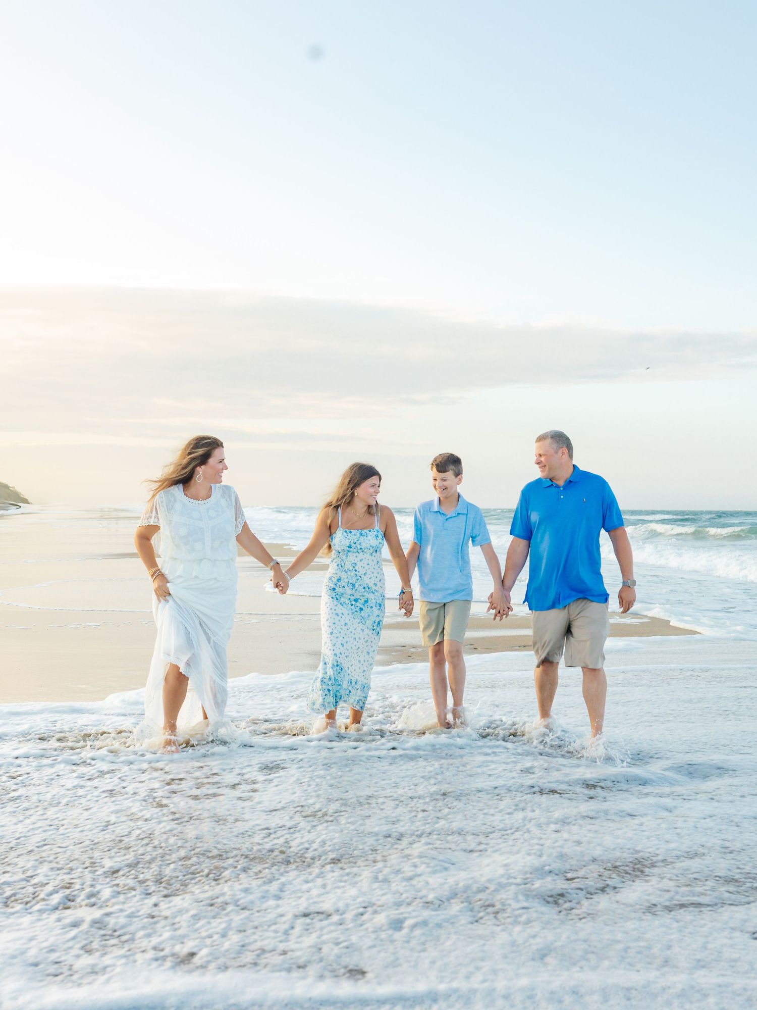 Family of four walking hand in hand along shoreline during sunset beach photoshoot.