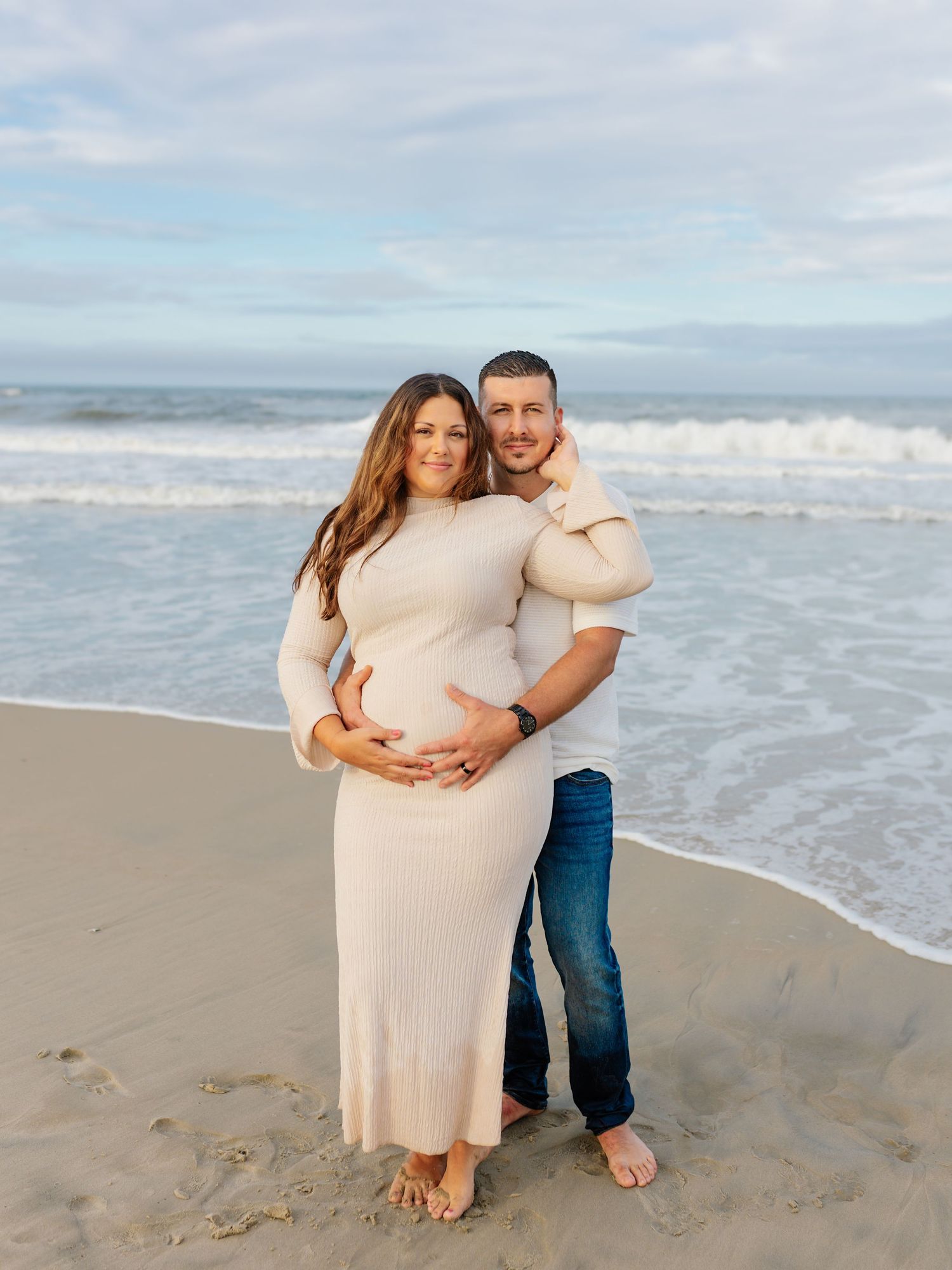 Romantic maternity photoshoot at the beach with couple in cream dress and casual attire.
