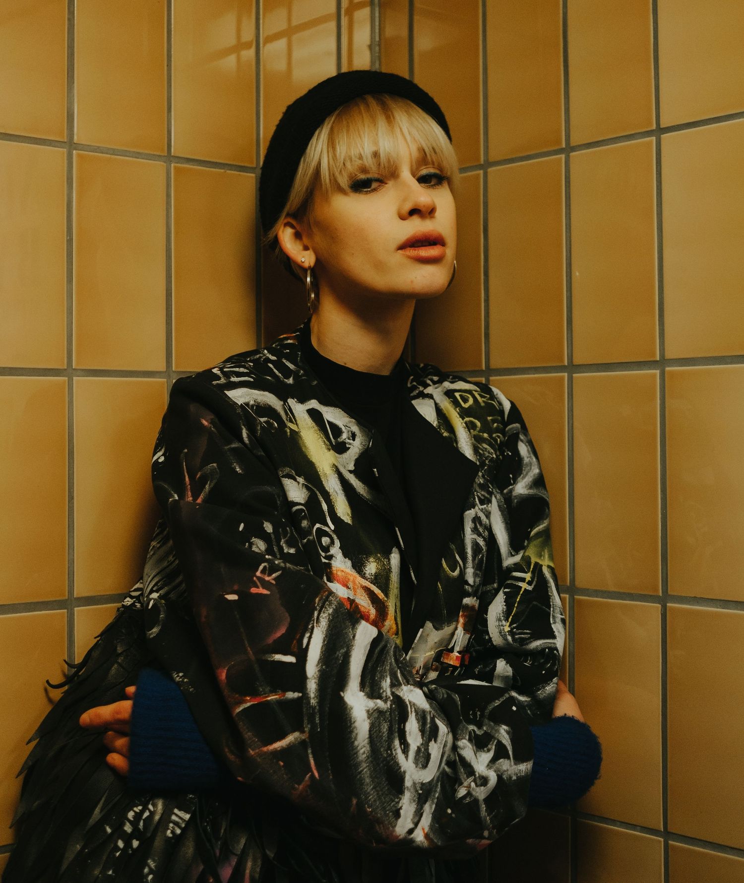 Person in black leather jacket poses against orange wall with metallic grid pattern in artistic photoshoot.