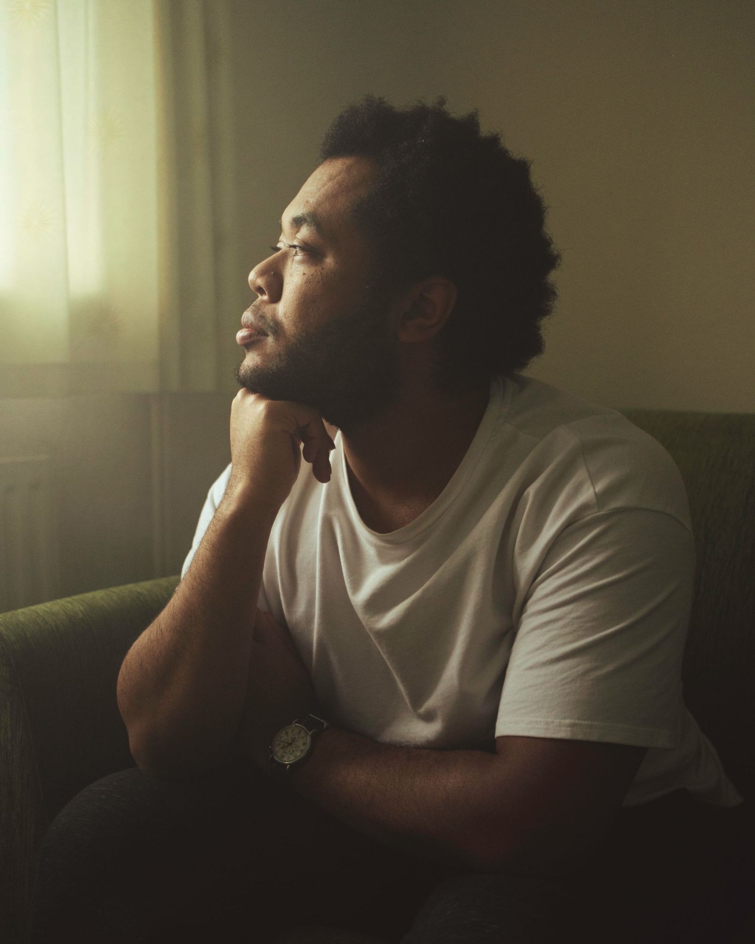 Person in white t-shirt sits in contemplative pose against soft window light creating moody atmospheric scene.