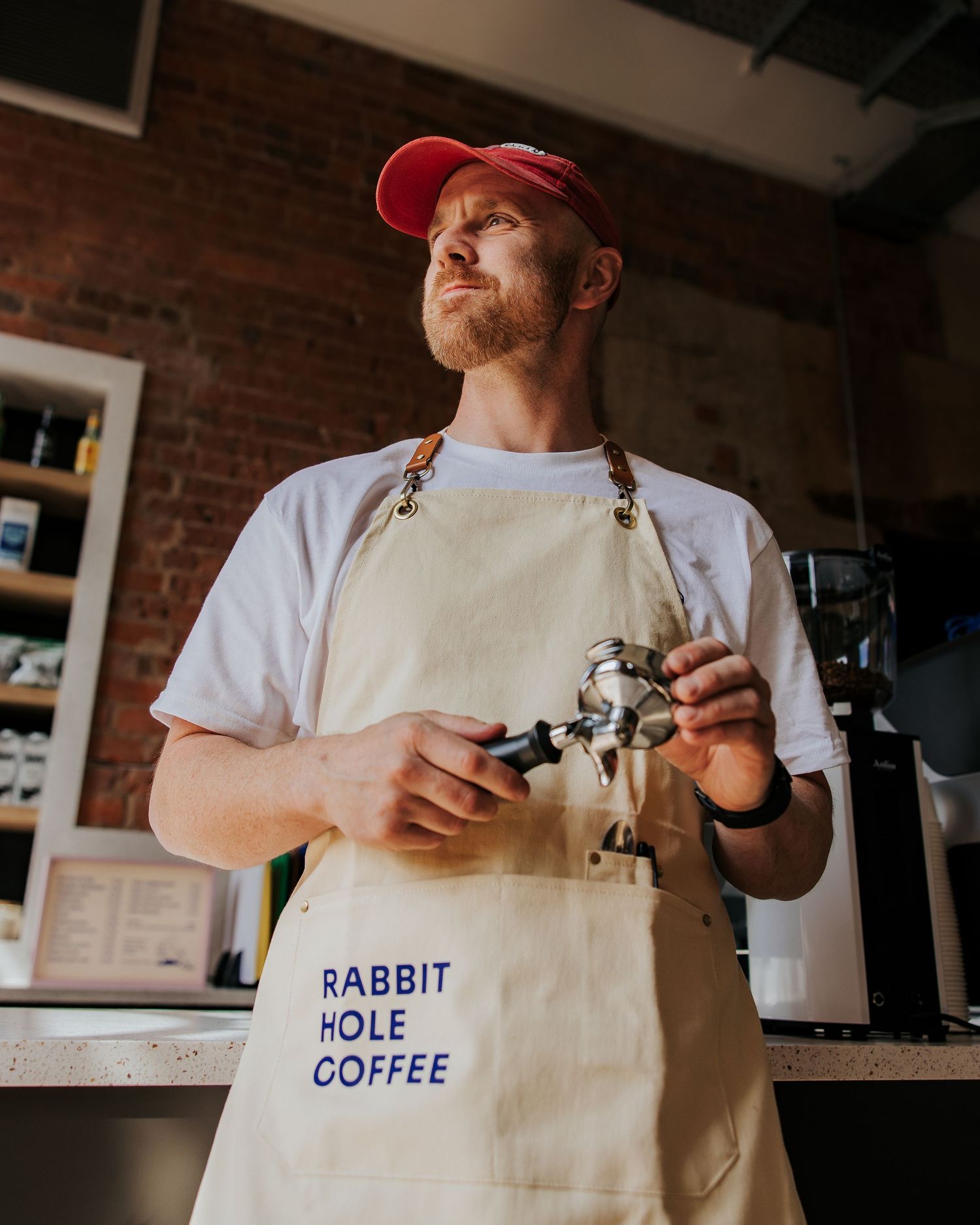 Barista wearing white t-shirt and beige apron with Rabbit Hole Coffee logo prepares drinks behind counter.