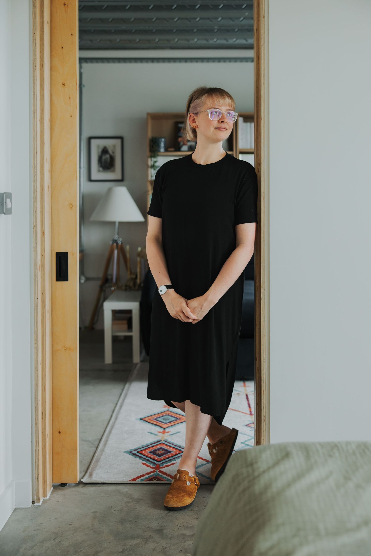 Someone in a black dress stands in a doorway with wooden trim and white walls, wearing brown leather shoes.