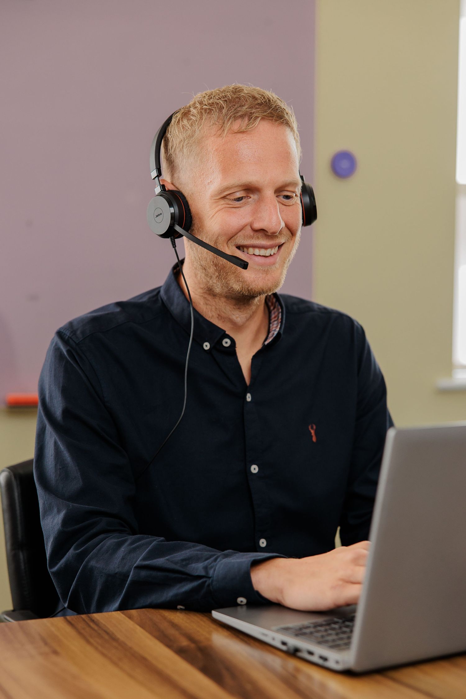 Customer service representative wearing headset smiles while working on laptop at wooden desk.