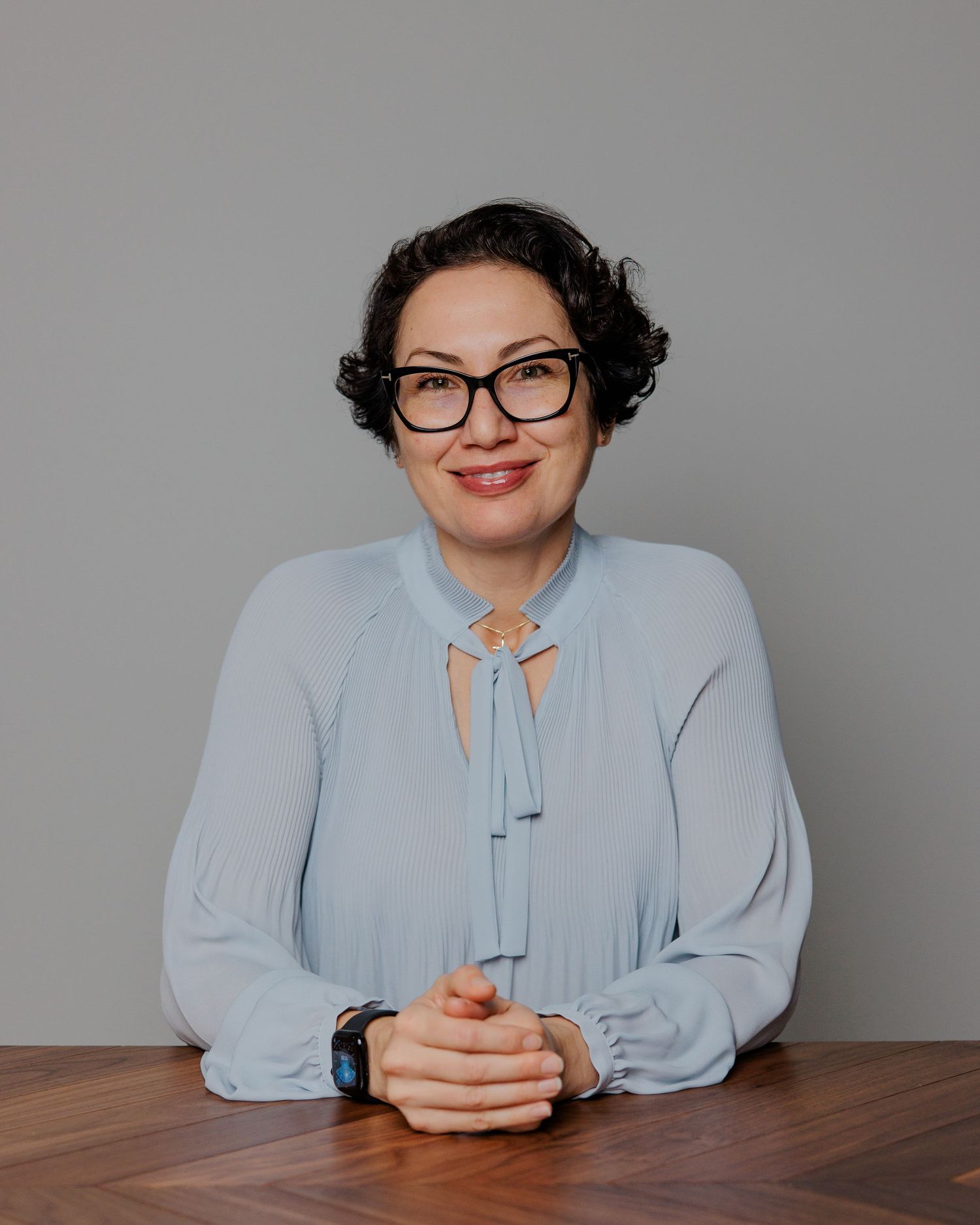A person in a light blue blouse and glasses sits at a wooden table in a professional studio setting.