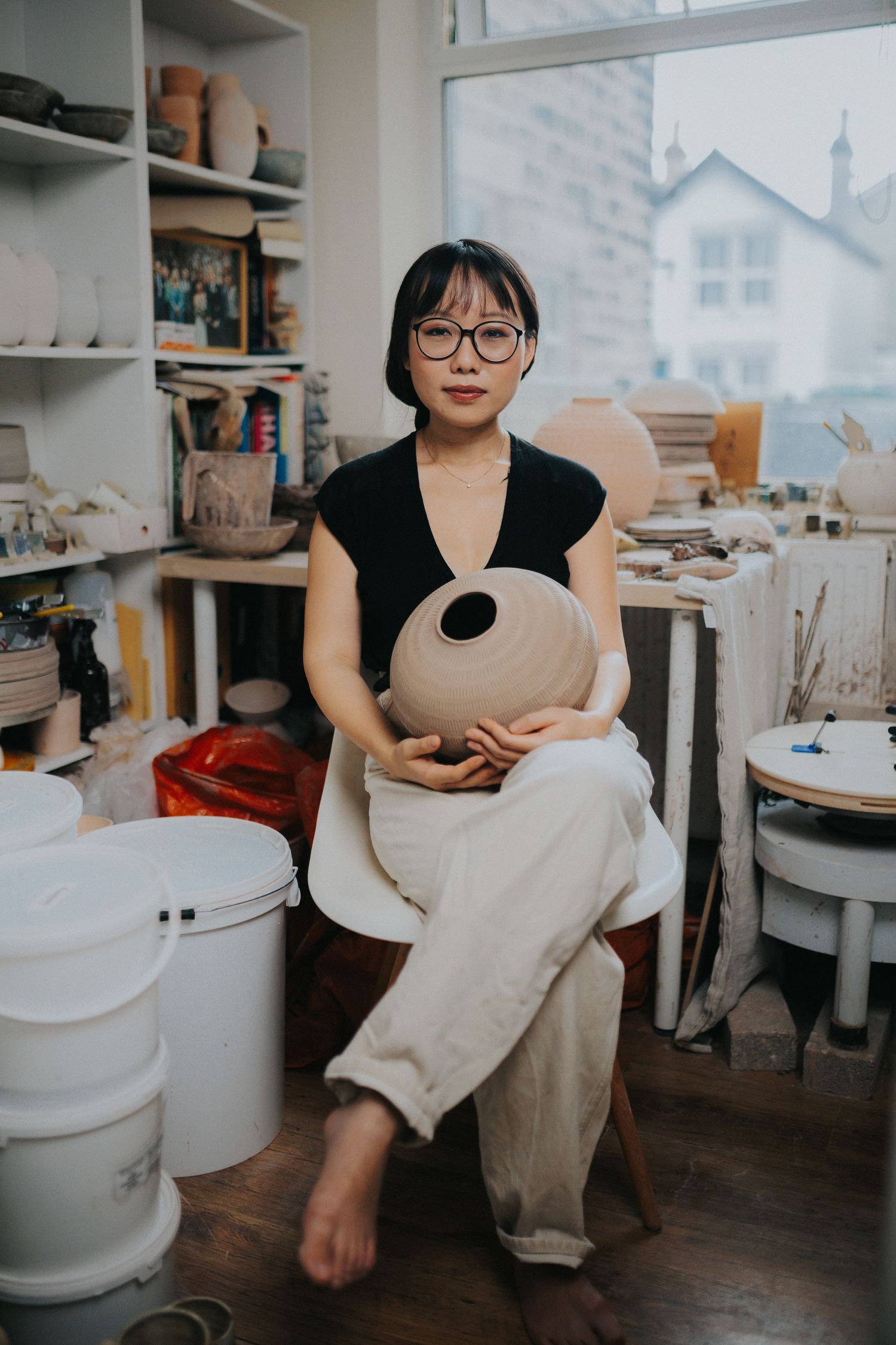 Artist working with clay pottery in a sunlit ceramic studio while sitting on a wooden stool.