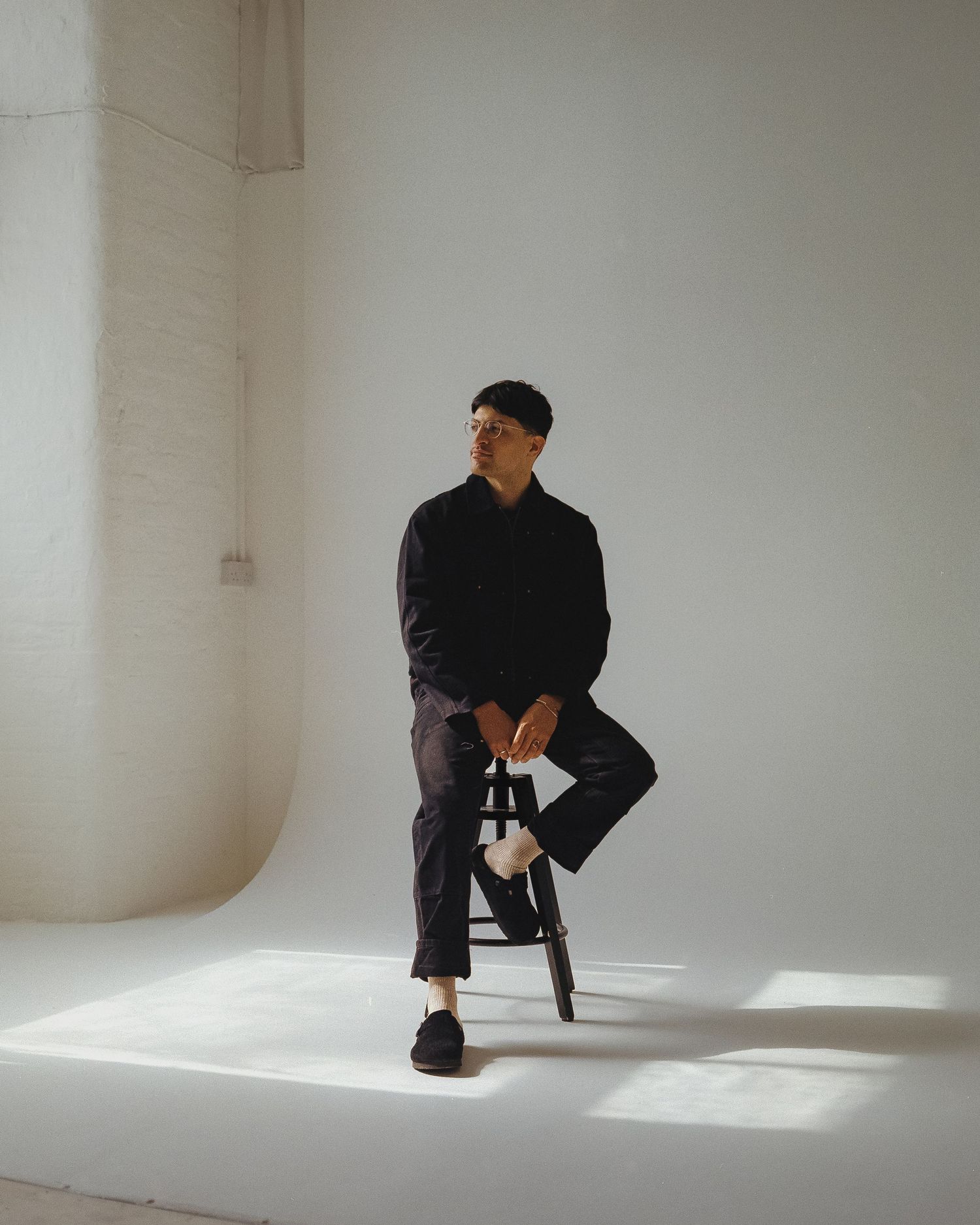 Minimalist fashion portrait in white room showing person in black outfit sitting on small stool with dramatic lighting.