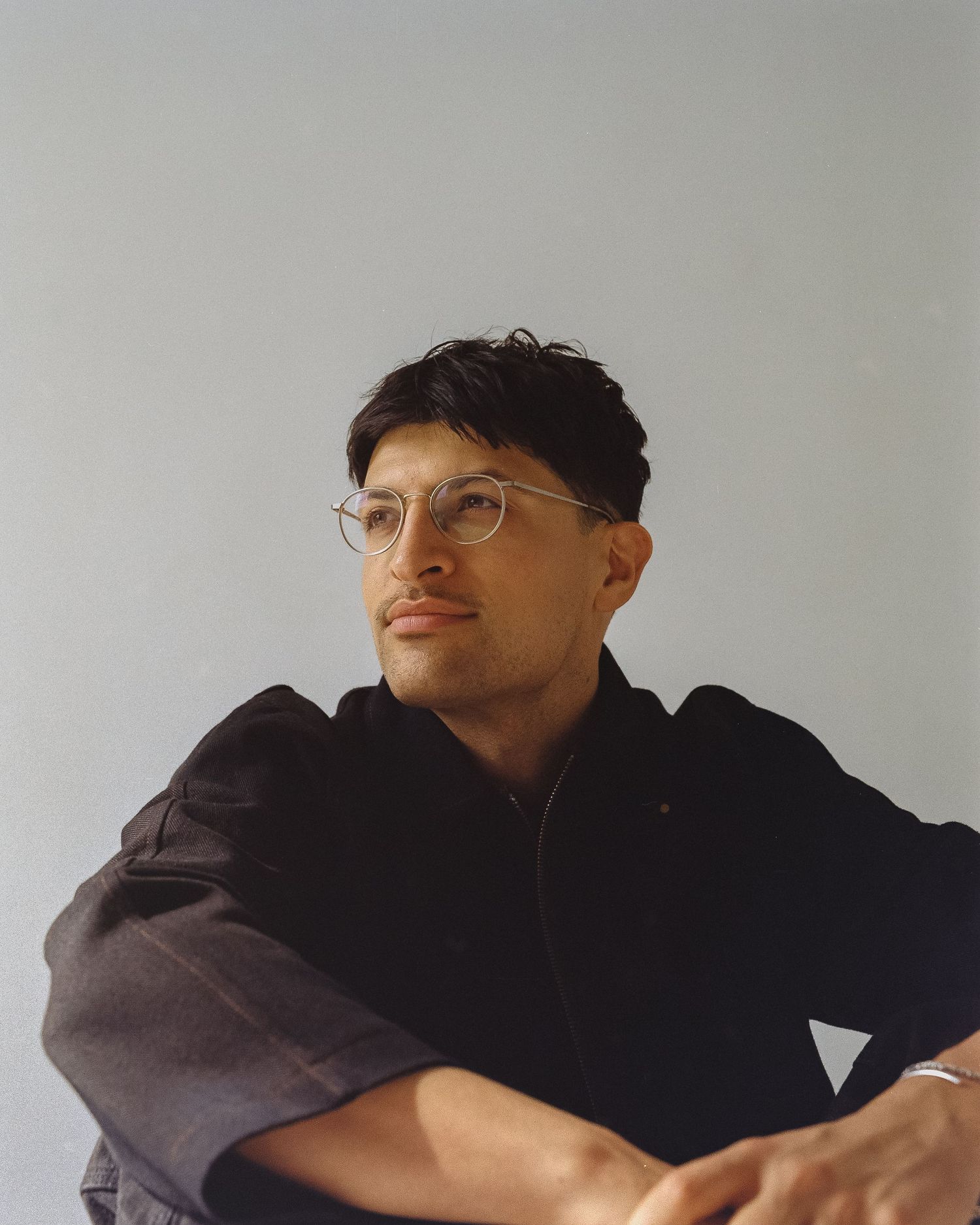A person in glasses and dark clothing poses against a light background in a minimalist studio setting.