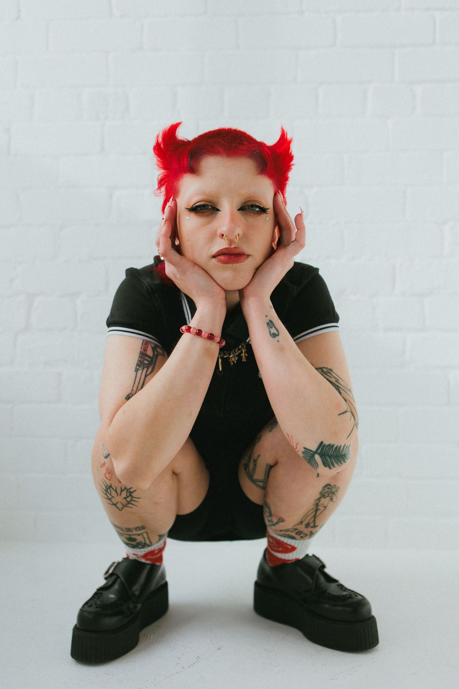 Person with bright red hair and tattoos crouches in casual black outfit against white wall.