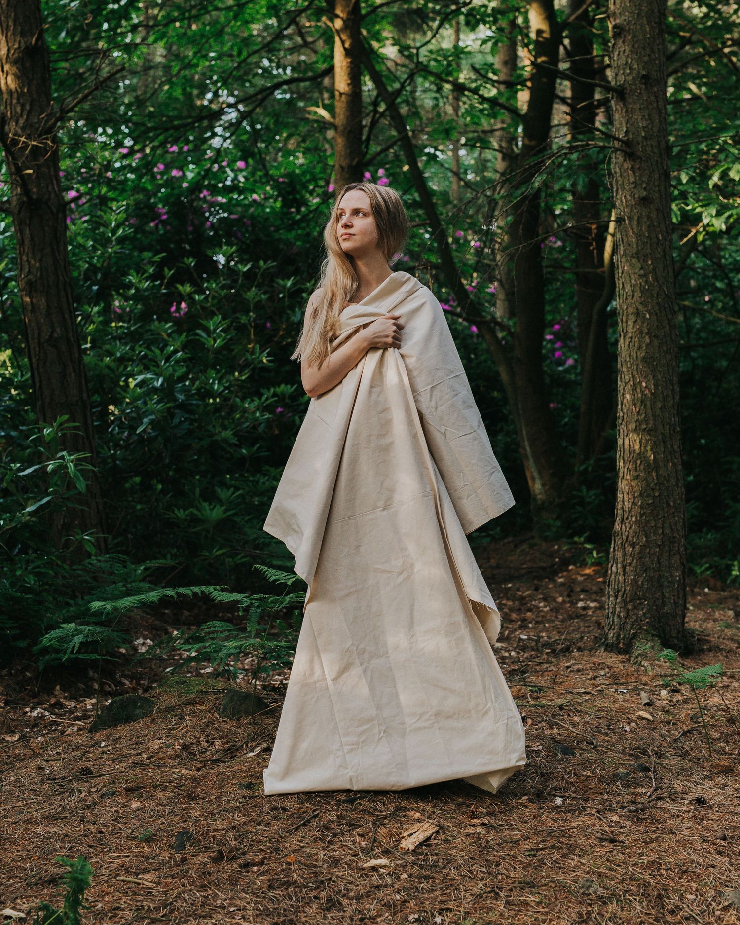 A person in a flowing beige dress stands contemplatively in a lush forest setting during golden hour.
