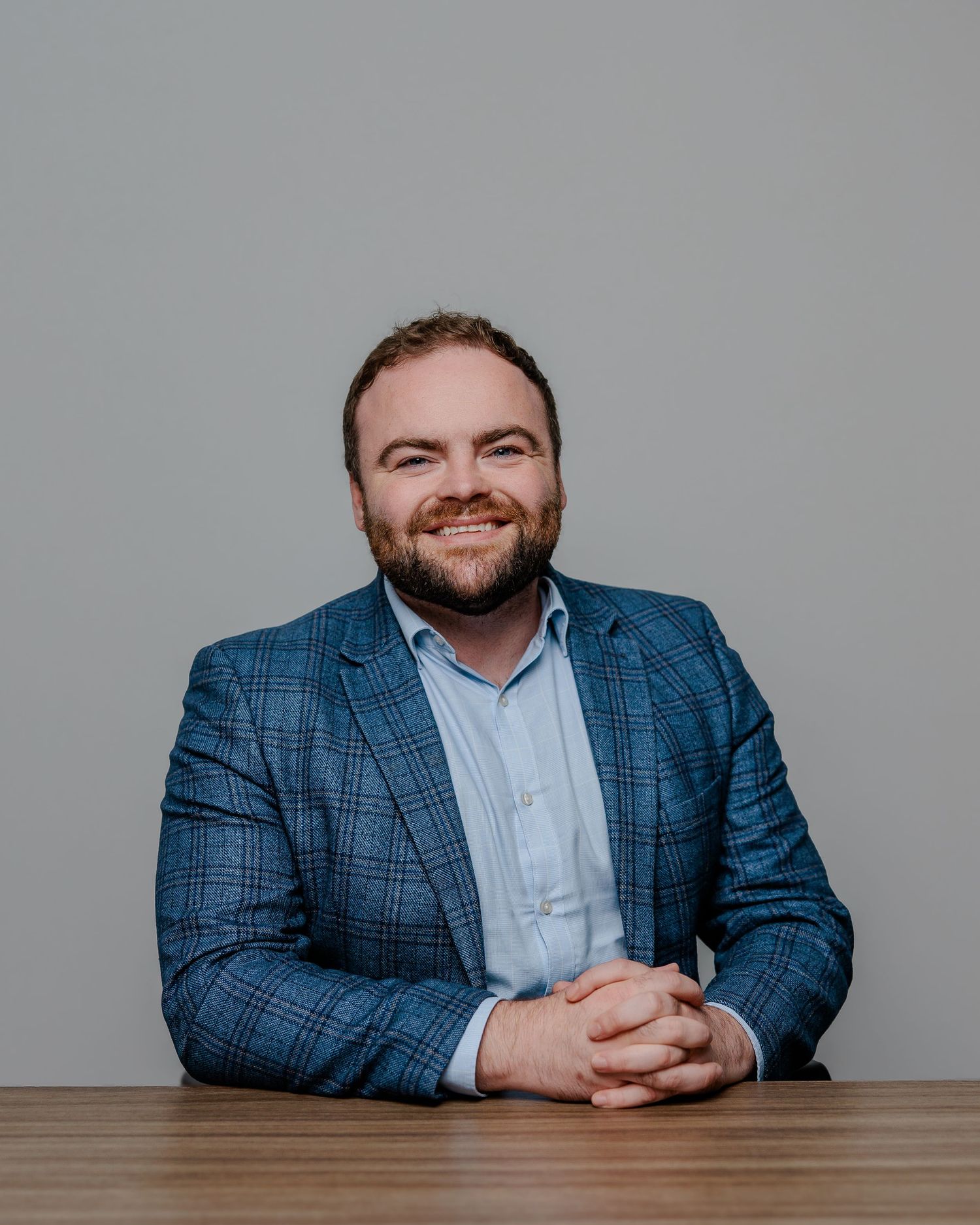 Professional headshot of someone wearing a blue plaid blazer and light blue shirt sitting at a wooden table.