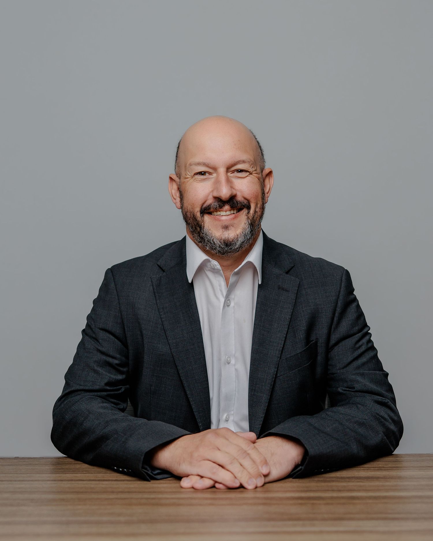 Professional portrait of a person in a dark suit and white shirt sitting at a wooden desk with a gray background.