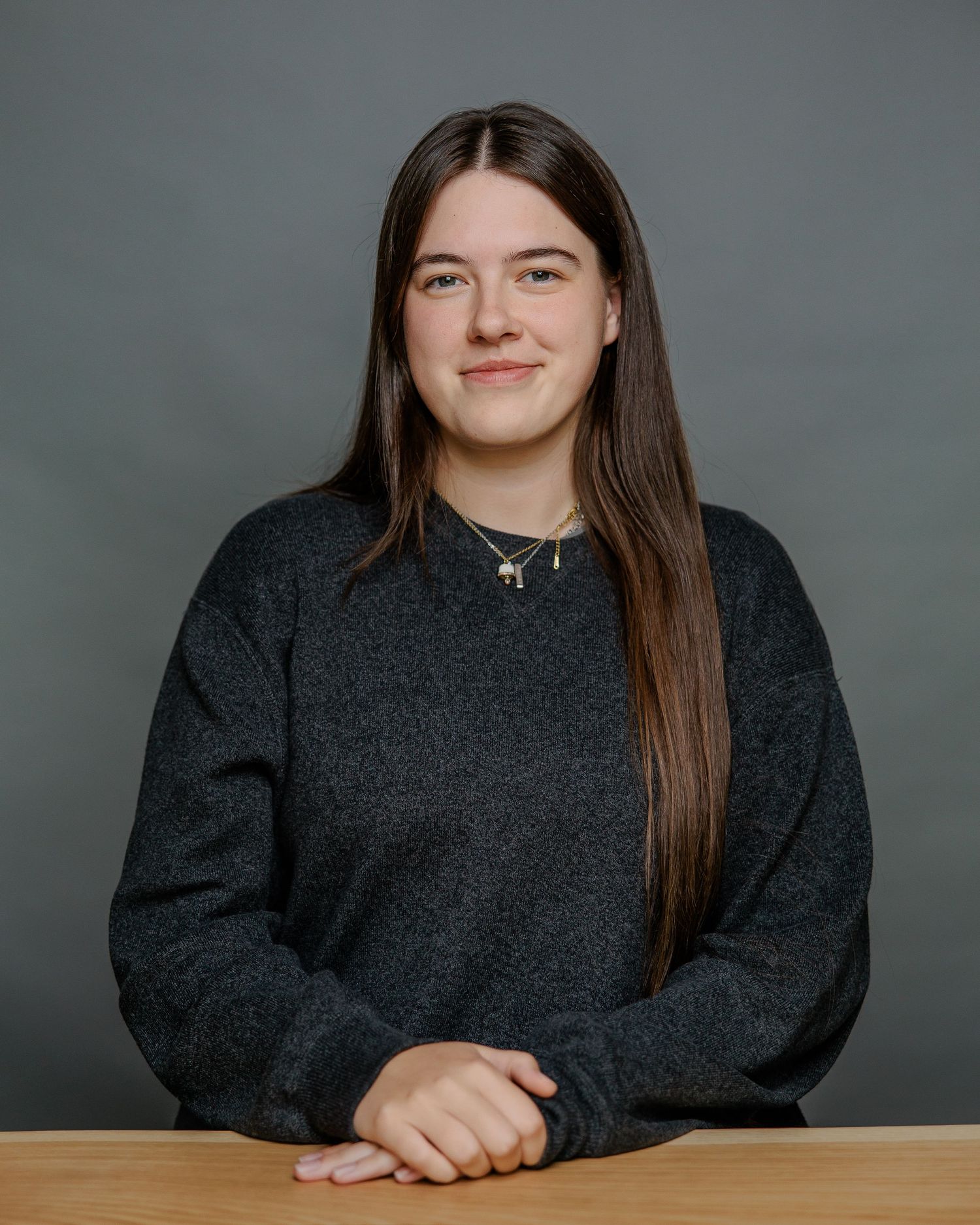 A person in a dark gray sweater sits at a desk against a gray background, showing a warm smile.