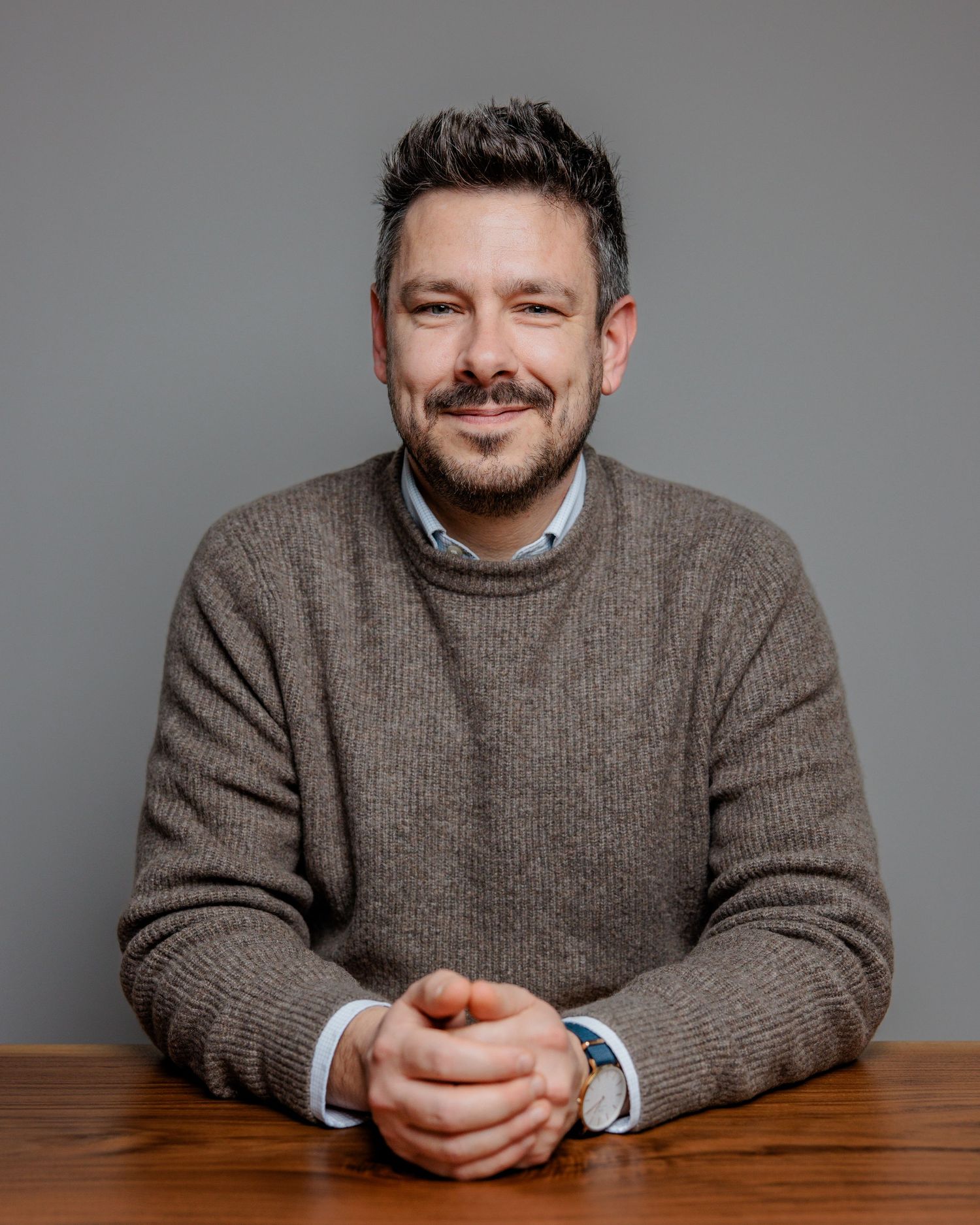 Person in a brown knit sweater leans on wooden table while smiling at the camera.