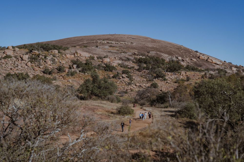 A panoramic view of a desert landscape with rocky hills, sparse vegetation, and hikers on a trail under blue skies.