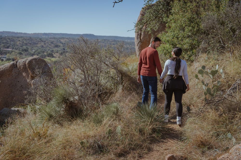 Two people hiking along a scenic mountain trail overlooking a vast desert canyon landscape.