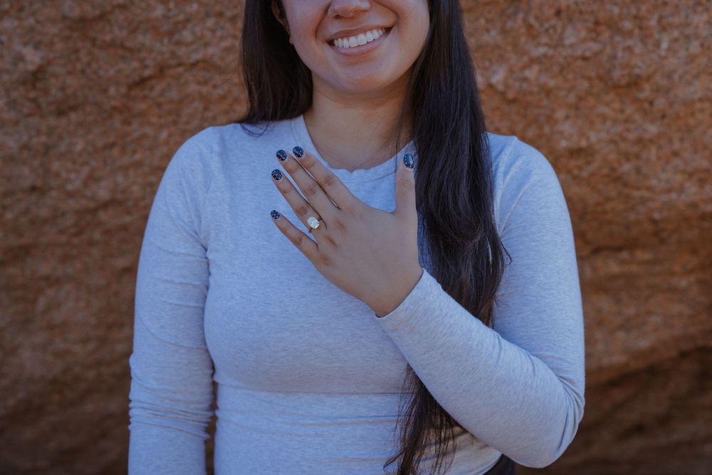 A person in a light gray long sleeve shirt smiles while standing against a textured brown wall.