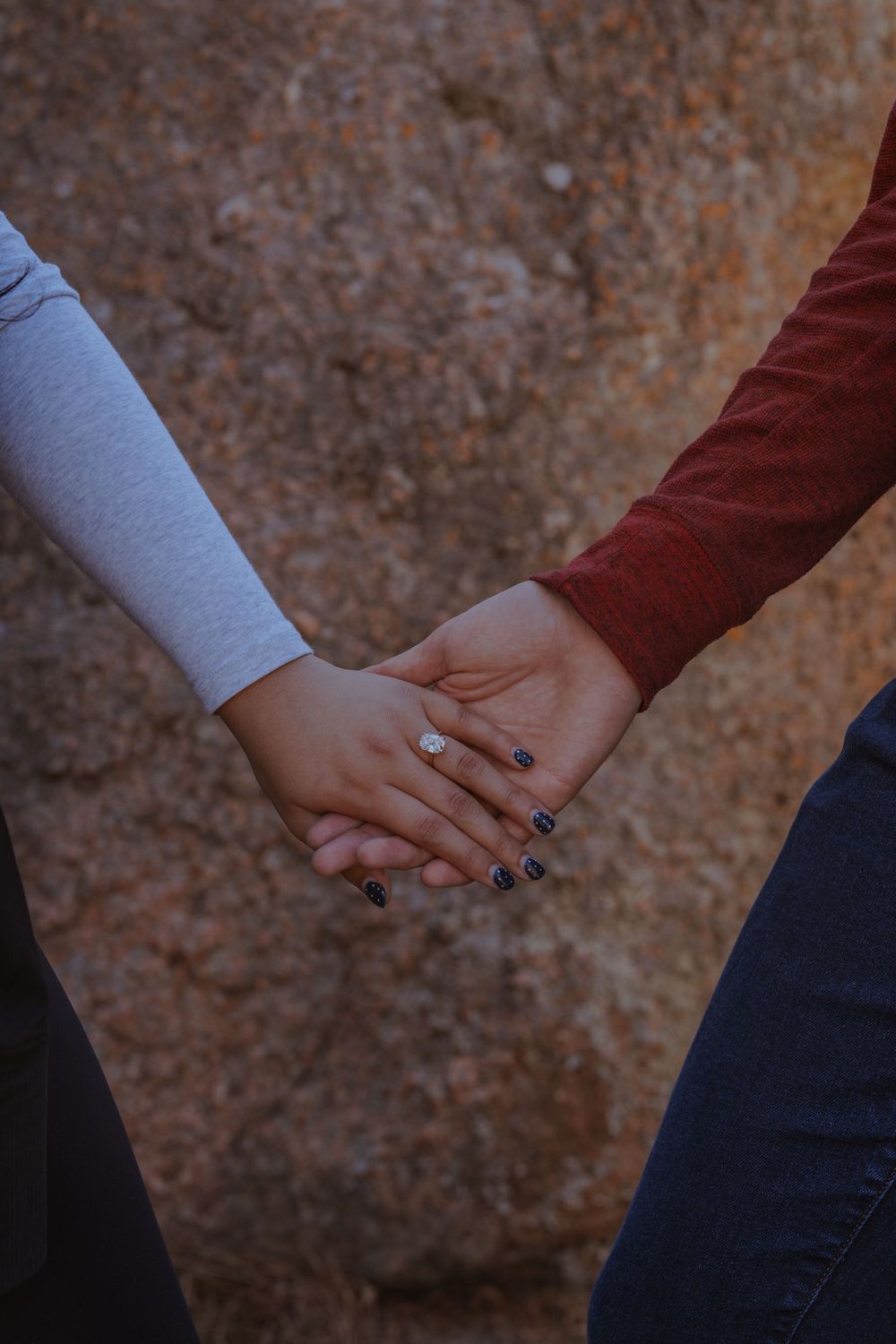 Two people holding hands with painted nails against a textured brown rock background.