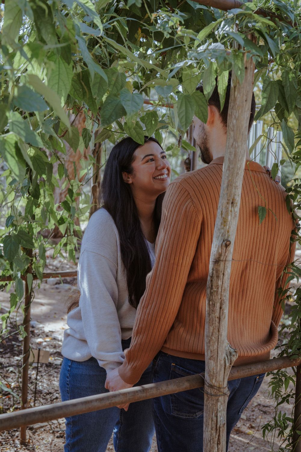 Two people sitting together on a wooden bench outdoors surrounded by green foliage on a sunny day.