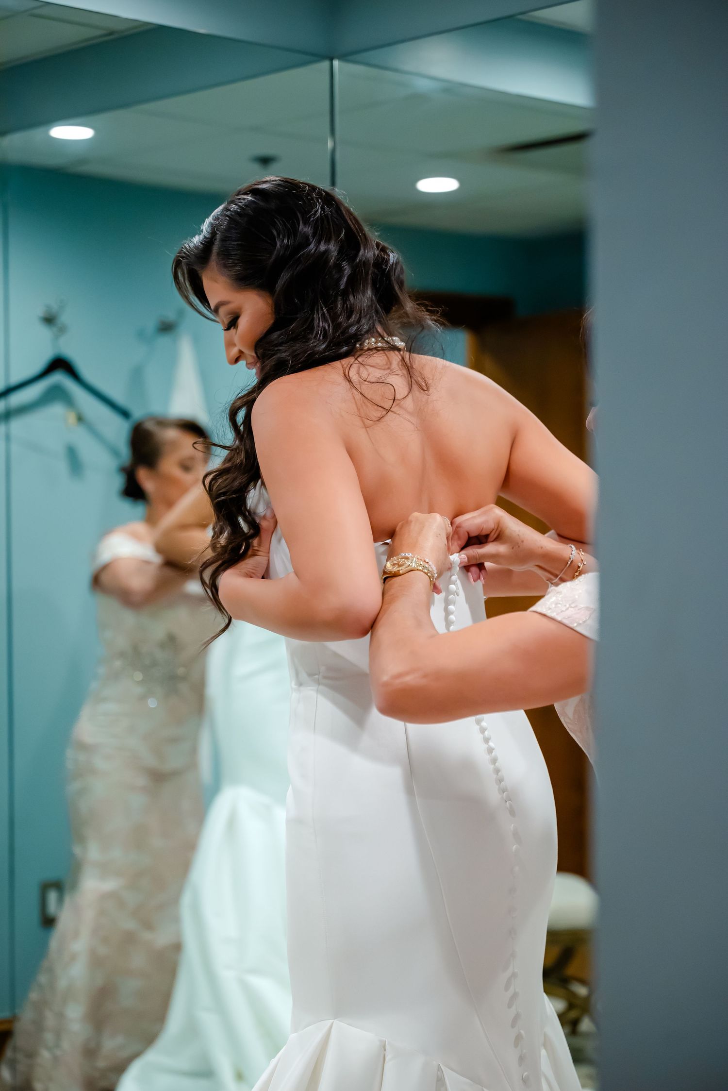 Woman in white wedding dress getting ready in dressing room with mirror reflection in background.