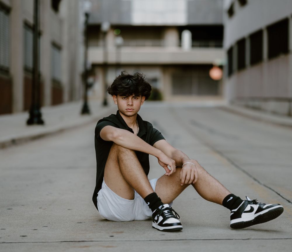 A casual urban portrait sitting on sidewalk wearing sneakers and shorts against city backdrop.